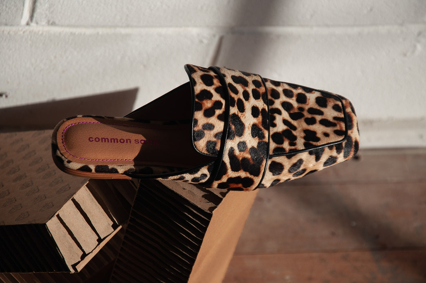 A right foot leopard print furry slip on mule with soft squared toe and black piping on edge. The mule is creatively placed on some corrugated cardboard. The background is a white painted brick wall and dusty floorboards.