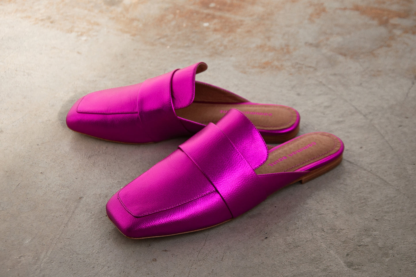 A pair of metallic pink slip on mules. The mule has a soft square toe and a wide band at top of foot. The mules are set on a concrete floor.