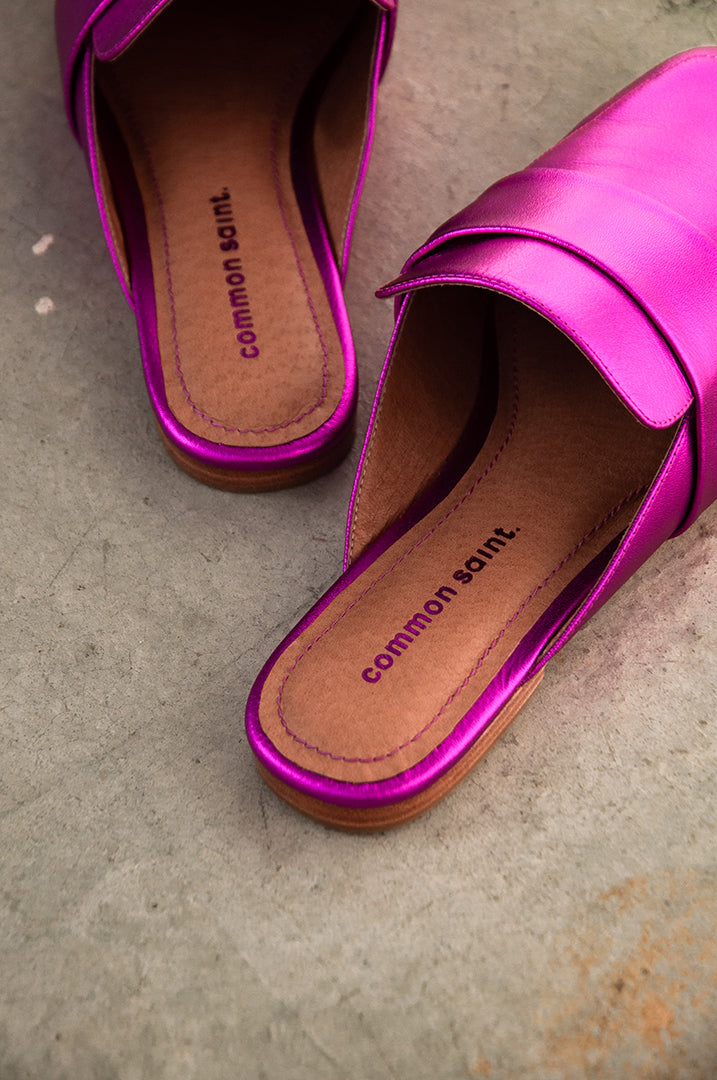 A pair of metallic pink lip on mules seen from the footbed. The brand Common Saint can be seen on footbed with matching pink stitching on the edge of the footbed. The mules are set on grey concrete flooring.