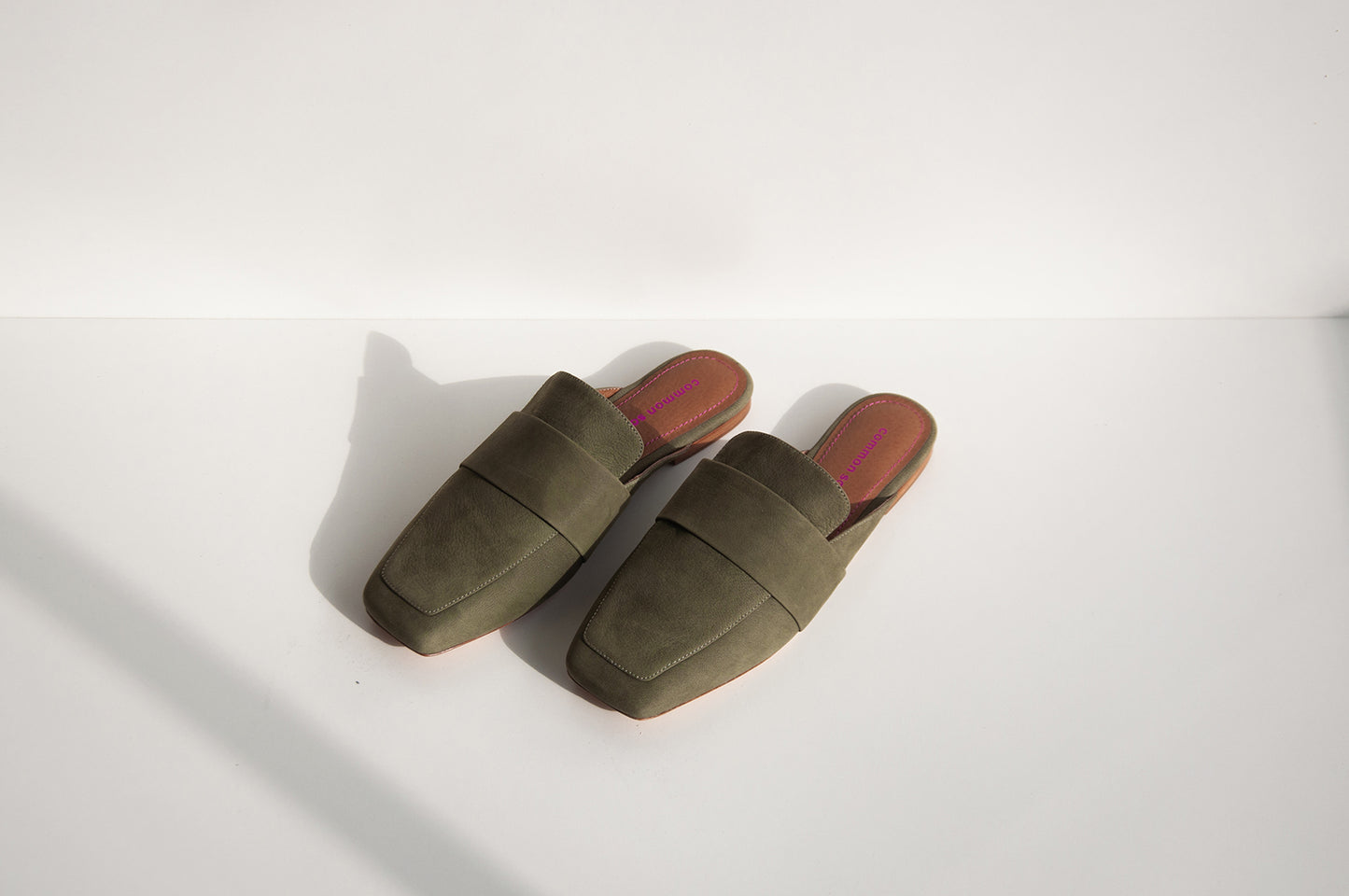 A pair of khaki mules on a white background with some shadow. The brand, Common Saint, can be seen on the footbed in metallic pink print and embossed. There is matching metallic pink stitching around the edges of the footbed.