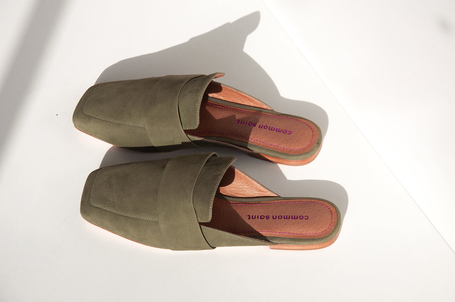 A pair of khaki slip on mules with a wide band. The shoes are placed on a horizontal angle. It is on a white background, with some shadows. THe brand common saint can be seen on the footbed in metallic pink and embossed. There is metallic pink stitching to match the branding along the edge of footbed.