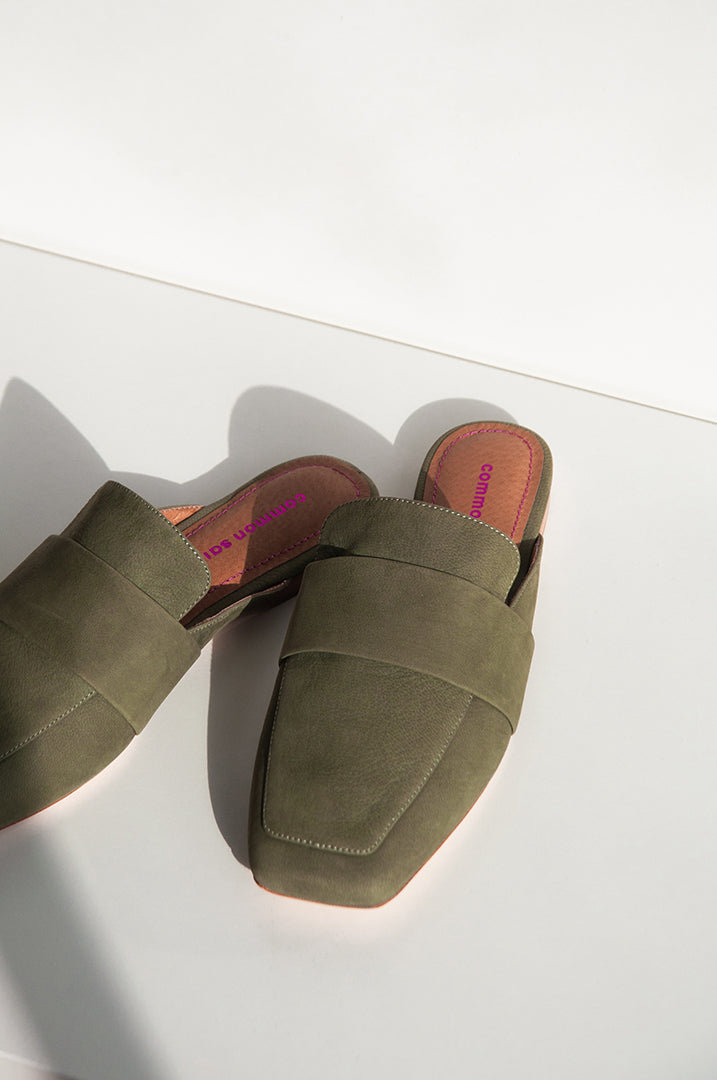 A pair of khaki leather mules with a soft square toe with some shadow. on a white background. Common Saint brand can be seen on the footbed in metallic pink colour and metallic pink stitching.