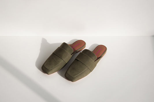 A pair of khaki mules on a white background with some shadow.  The brand, Common Saint, can be seen on the footbed in metallic pink print and embossed.  There is matching metallic pink stitching around the edges of the footbed.