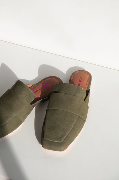 A pair of khaki leather mules with a soft square toe with some shadow. on a white background.  Common Saint brand can be seen on the footbed in metallic pink colour and metallic pink stitching.